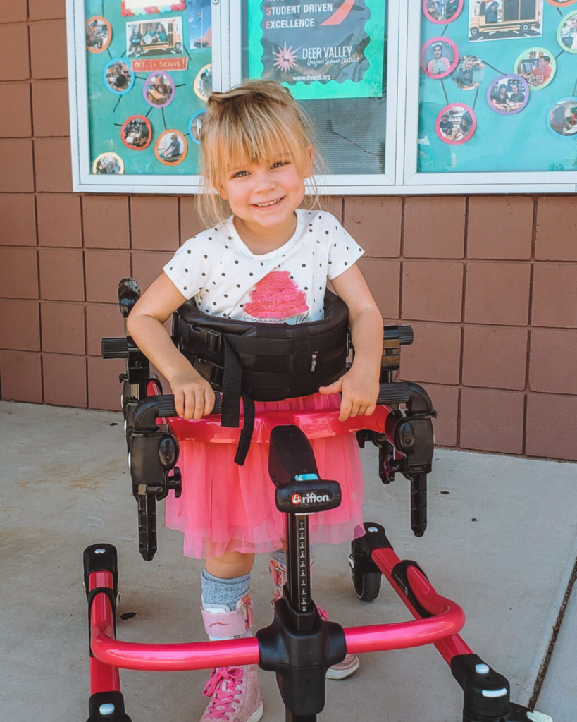 A young girl with blond hair smiles while using a pink walker. She wears a white polka dot shirt, a pink tutu, and pink shoes. Behind her is a brick wall with colorful posters and photos.