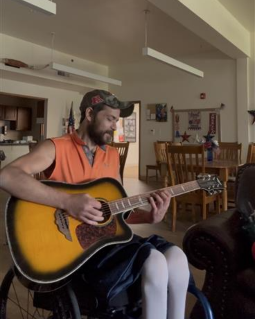 A man in a sleeveless orange shirt and cap sits in a wheelchair playing an acoustic guitar in a cozy, well-lit room with wooden furniture and patriotic decorations.