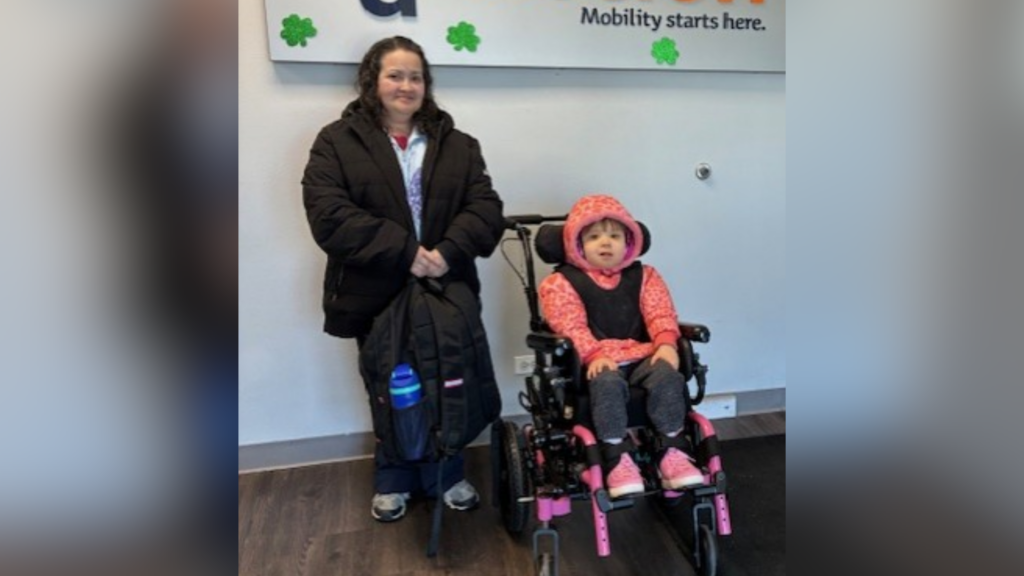 A woman in a black coat stands next to a young girl in a pink wheelchair and hooded jacket. Indoors, they pose in front of a wall with green shamrock decorations, sharing a moment inspired by Violet's Story and a motivational sign.