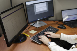 A person wearing a white sweater types on a keyboard at a desk with two computer monitors displaying ATP Development Program spreadsheets and forms, and a laptop, with a yellow highlighter and sticky notes nearby.