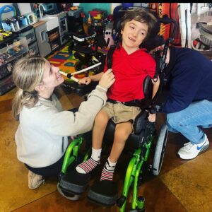 A smiling boy in a red shirt and tan shorts sits in a green wheelchair. Two adults, part of the ATP Development Program, kneel beside him, helping with his straps. Toys and colorful play mats are visible in the background.
