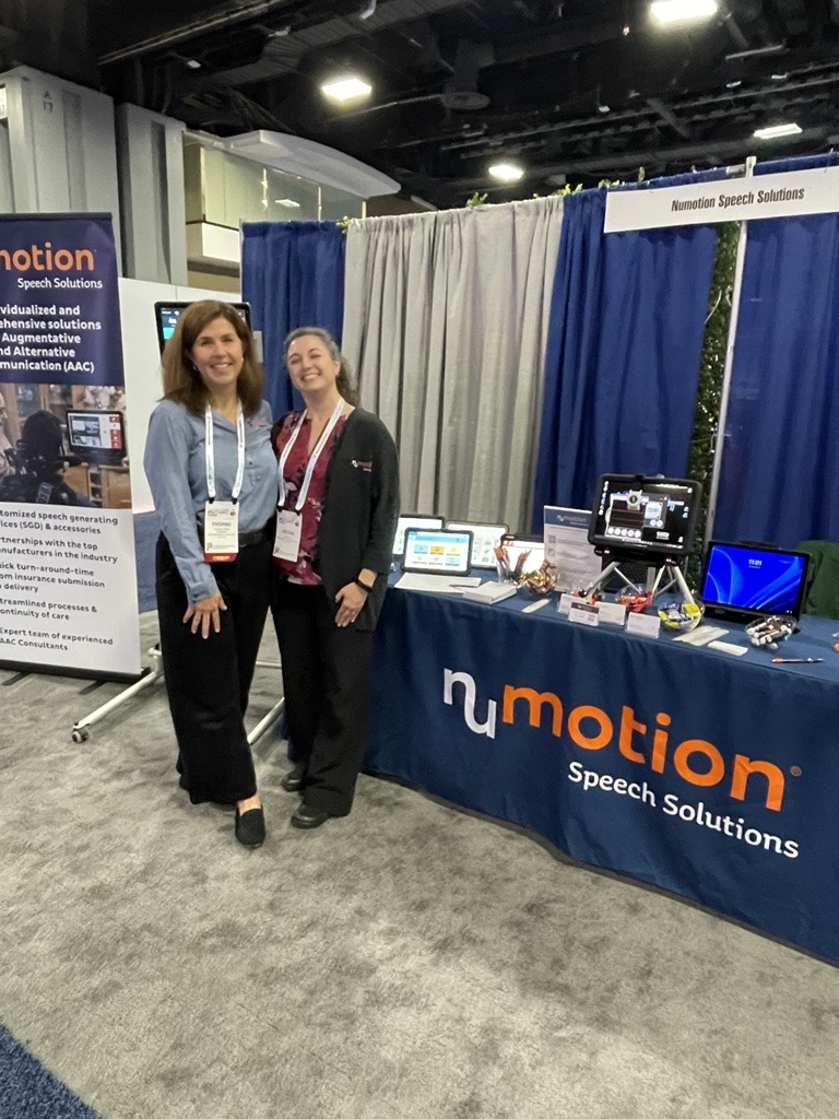 Two women stand smiling in front of a booth for Numotion Speech Solutions at a convention, where Yvonne's Story is featured alongside electronic devices, brochures, a display banner, and a blue tablecloth with the company logo.