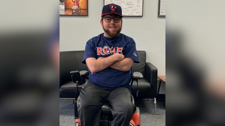 A man with a beard and glasses sits in a wheelchair, smiling. He is wearing a navy blue ROME t-shirt and matching hat. Behind him are a black couch and framed photos, capturing the warmth of Carson's Story.