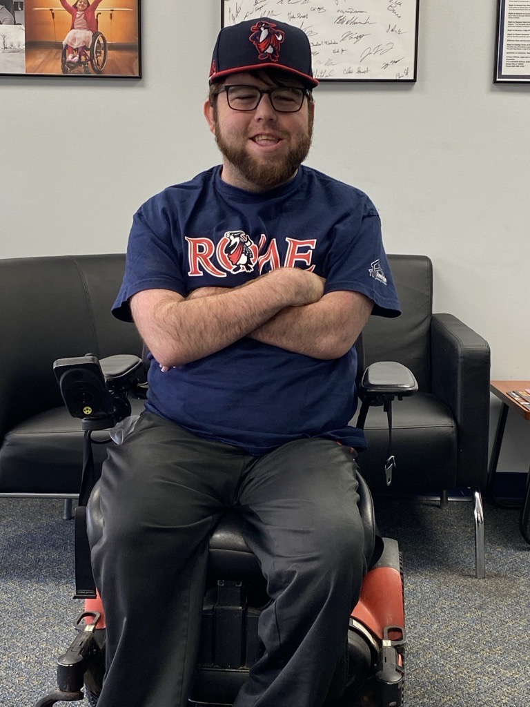 A man wearing glasses and a “ROME” t-shirt sits in a motorized wheelchair with arms crossed, smiling. He also wears a baseball cap. Behind him are a black sofa and framed posters—a cozy glimpse into Carson's Story.