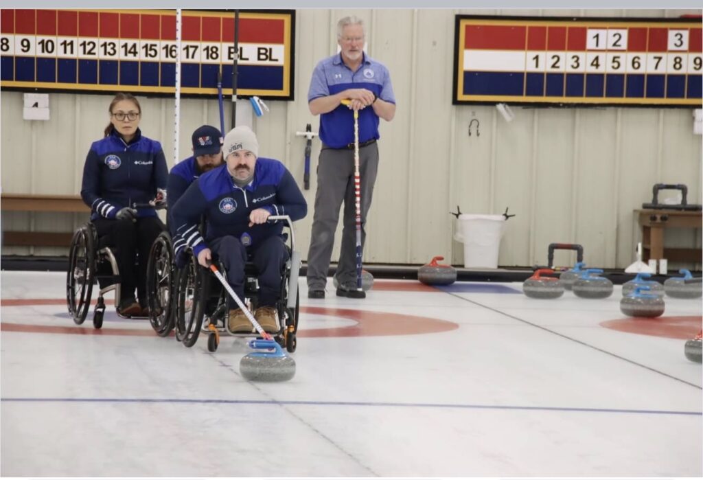Four people are curling on an indoor ice rink. In Sean's Story, two team members in wheelchairs launch a stone, a third teammate observes, and their coach stands behind them. Curling stones and scoring boards are visible in the background.