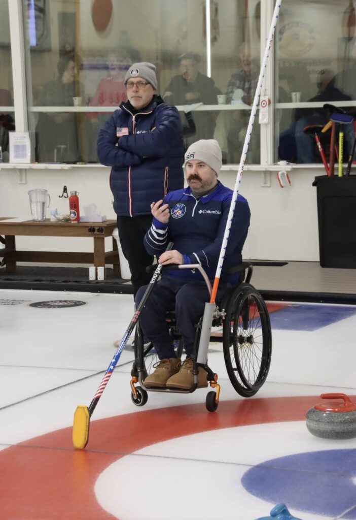 A man in a wheelchair prepares to throw a curling stone on an ice rink, holding a delivery stick, while another man stands behind him watching. Both are wearing winter clothing and beanies, capturing a moment from Sean's Story.