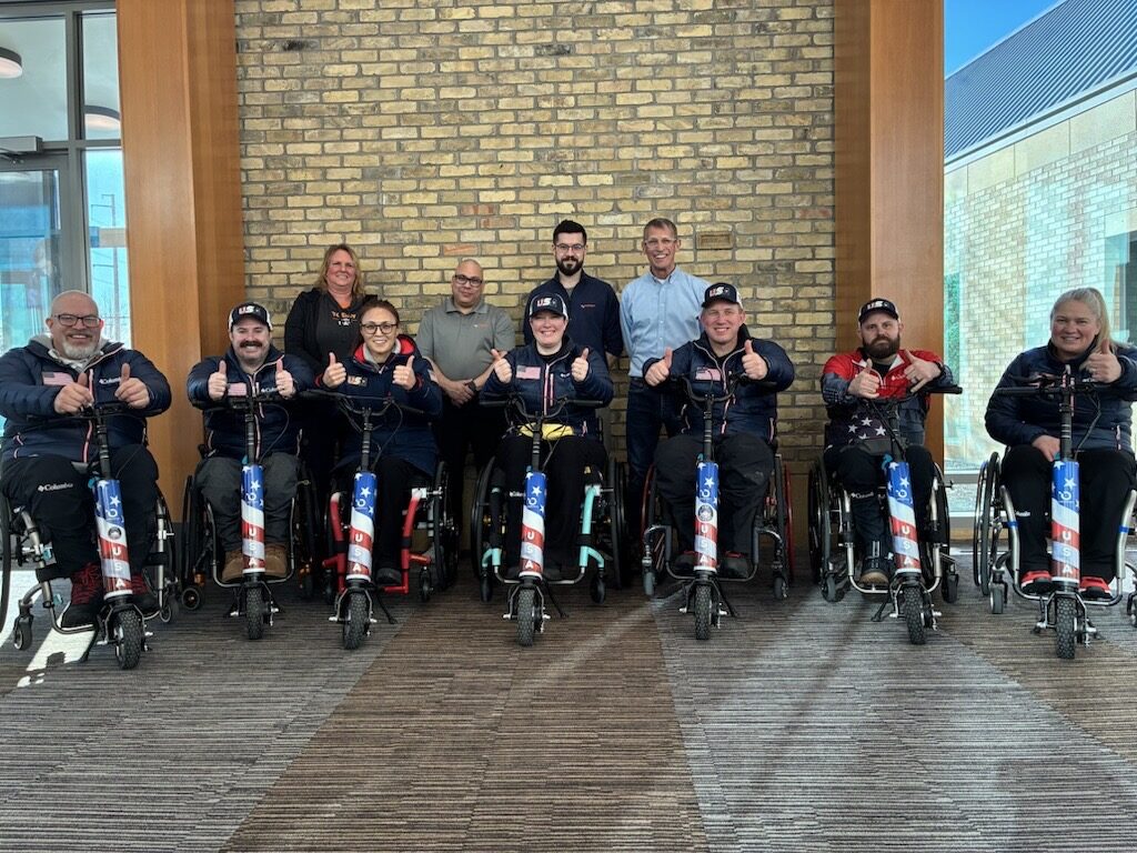A group of ten people, some in wheelchairs decorated with American flag designs, pose indoors, smiling and giving thumbs up in front of a brick wall—capturing the spirit of Sean's Story. Four people stand behind six seated team members.