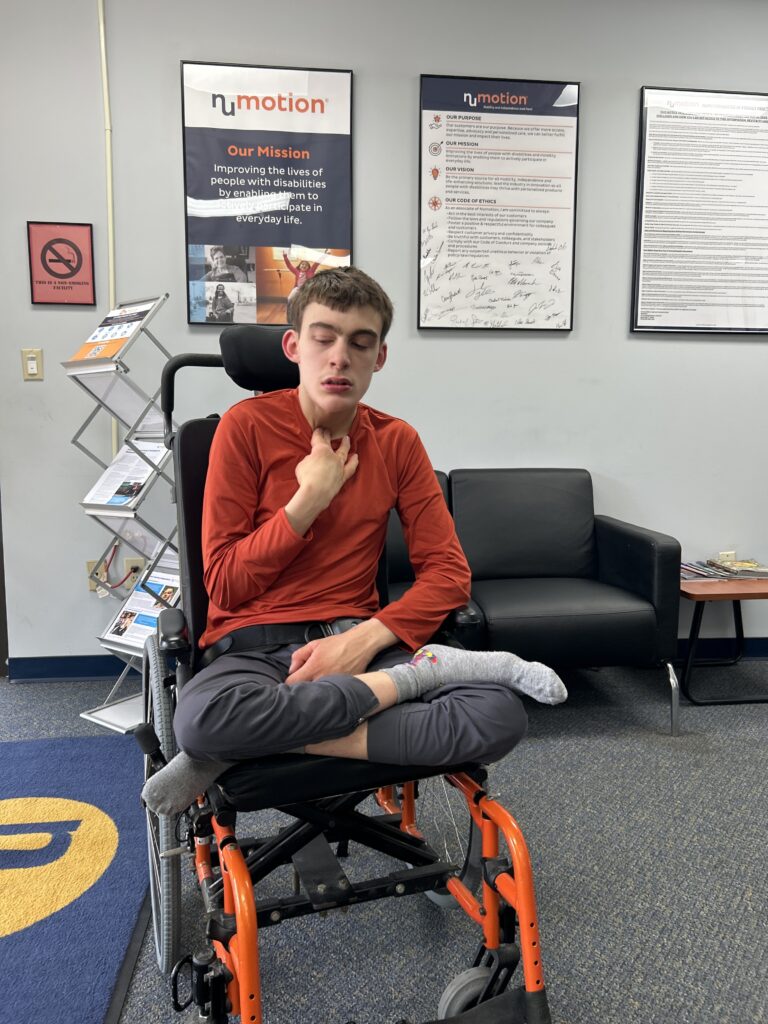 A young person in a red shirt and gray pants sits in an orange wheelchair, eyes closed, with one hand touching their chest. Behind them are informational posters and brochures sharing Jackson's Story in an office setting.