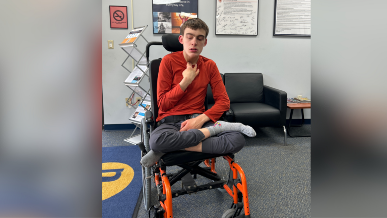 Jackson, a young person in a red long-sleeve shirt and gray pants, sits cross-legged in an orange wheelchair, touching their neck. Their story unfolds in a waiting area with black chairs, posters on the wall, and a pamphlet rack.