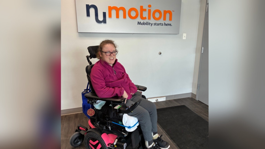 A woman in a pink jacket smiles while sitting in a power wheelchair in front of a wall sign that reads “numotion Mobility starts here,” capturing the uplifting spirit of Kelly's Story at an office or clinic entrance.