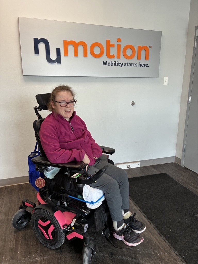 A smiling woman in a pink jacket sits in a power wheelchair with pink accents in front of a NuMotion sign that reads "Mobility starts here," capturing Kelly's Story in what appears to be an office or clinic lobby.