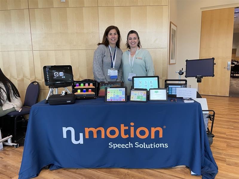 Two women stand behind a table covered with a blue NuMotion Speech Solutions tablecloth, displaying various speech device tablets and communication aids in a conference room, sharing Yvonne's Story of empowerment through communication.