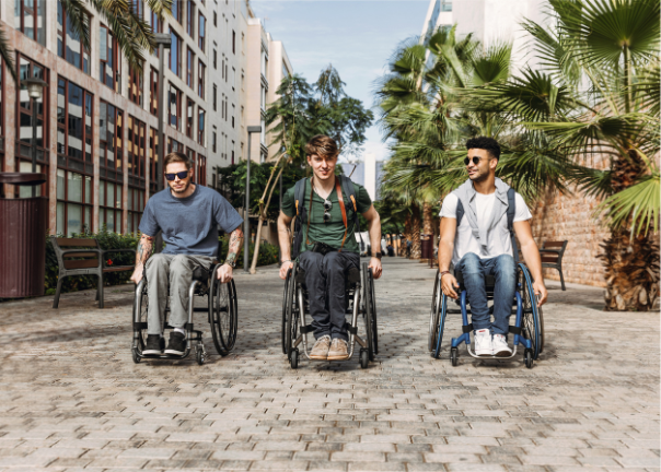 Three young men in wheelchairs, wearing casual clothes and backpacks, move along a sunny, tree-lined city street paved with bricks, smiling and enjoying each other's company—showing exactly what to expect from a day out with friends.