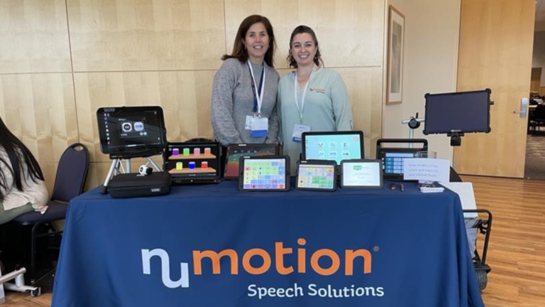 Two women stand behind a table displaying speech devices and tablets, with a blue tablecloth that reads NuMotion Speech Solutions, sharing Yvonne's Story in a room with light wood walls and framed artwork.