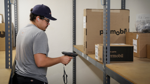 A man wearing a cap and glasses scans a cardboard box on a metal shelf in a storage room, stocking parts for repairs using a handheld barcode scanner.