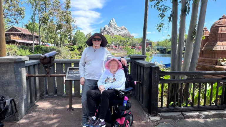 Two people pose together by a railing at a theme park for Terri's Story, one standing and one seated in a motorized wheelchair. They wear sun hats and sunglasses, with trees, water, and a mountain-shaped ride visible under the blue sky.