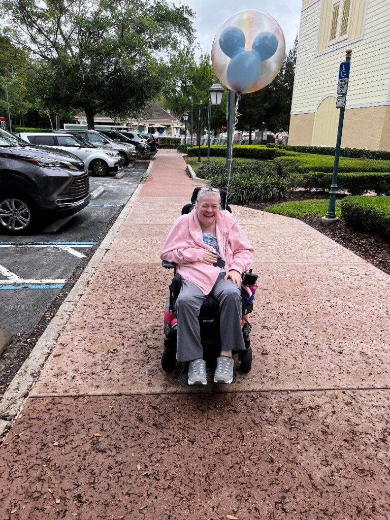 An older woman in a pink jacket sits in a motorized wheelchair on a walkway, smiling and holding a balloon with Mickey Mouse ears. Cars and greenery are in the background.