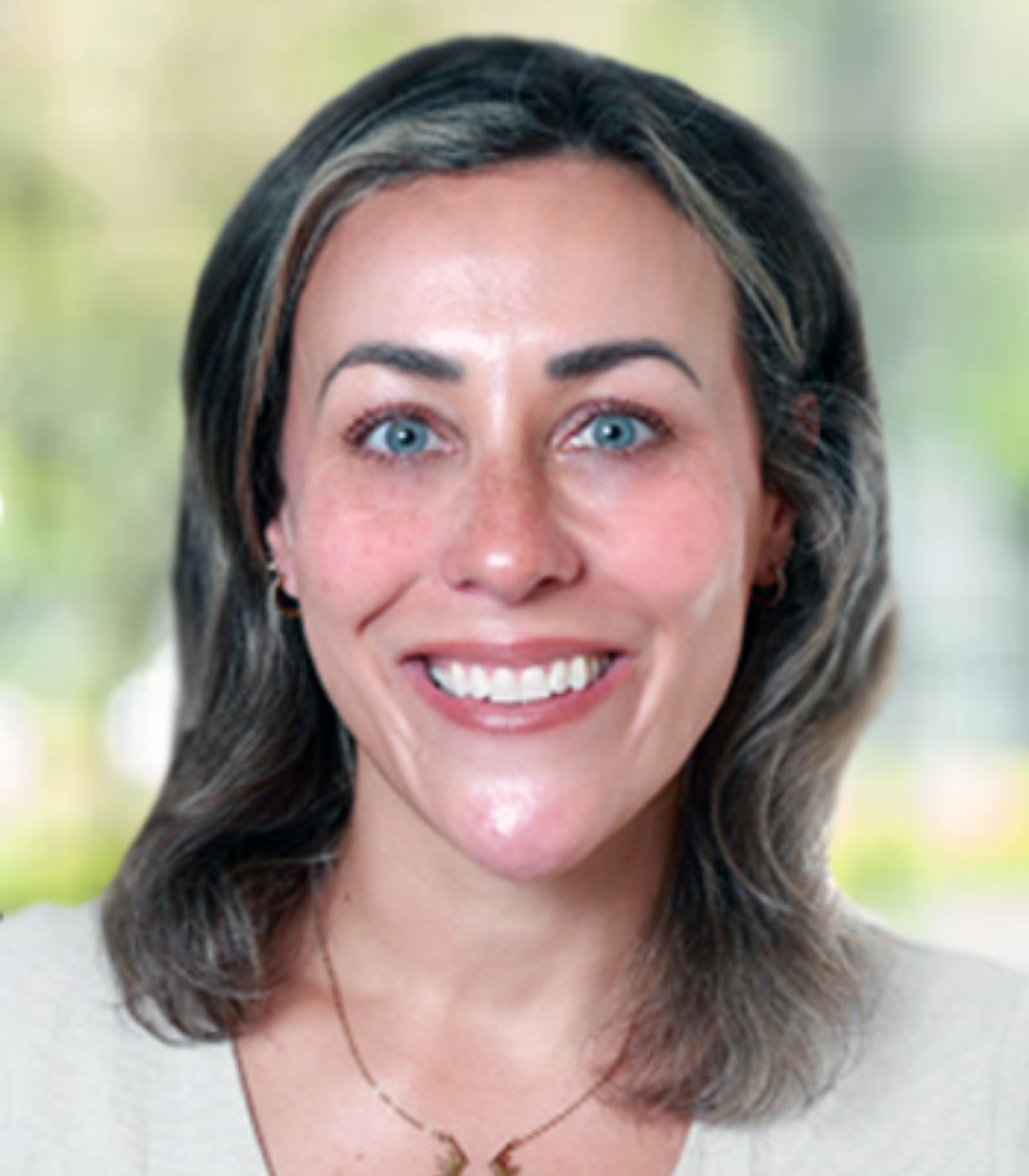 A woman with shoulder-length brown hair, blue eyes, and light skin smiles at the camera. She is wearing a light-colored top and a necklace, representing Speech Solutions coverage in a blurred outdoor background.