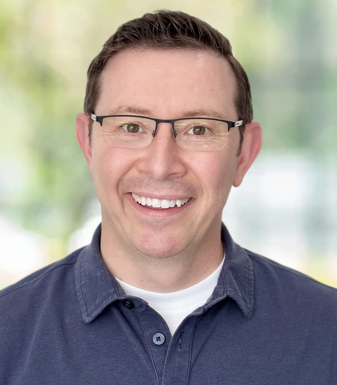 A man with short brown hair and glasses smiles at the camera. He is wearing a navy blue collared shirt over a white t-shirt. The background, softly lit with greenery, suggests he’s ready to discuss Speech Solutions or broaden coverage.